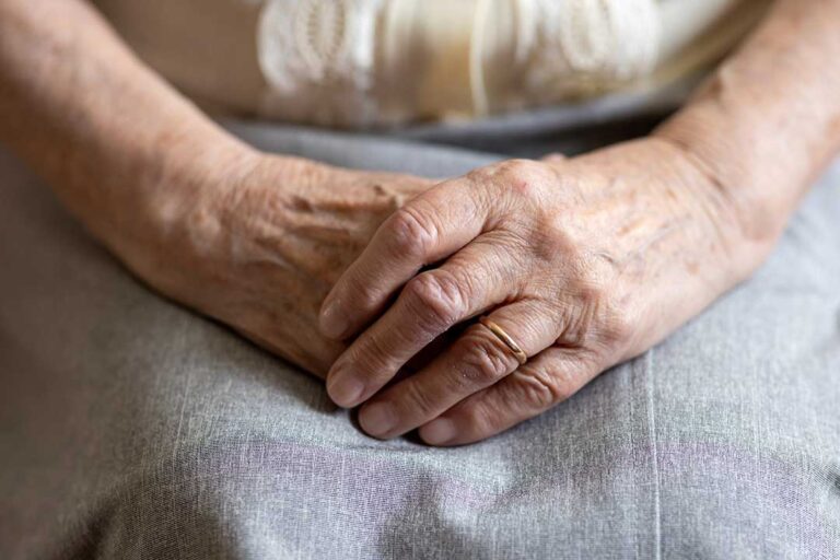 A caregiver hands medication to an older adult sitting at a kitchen table, both looking attentive and calm.