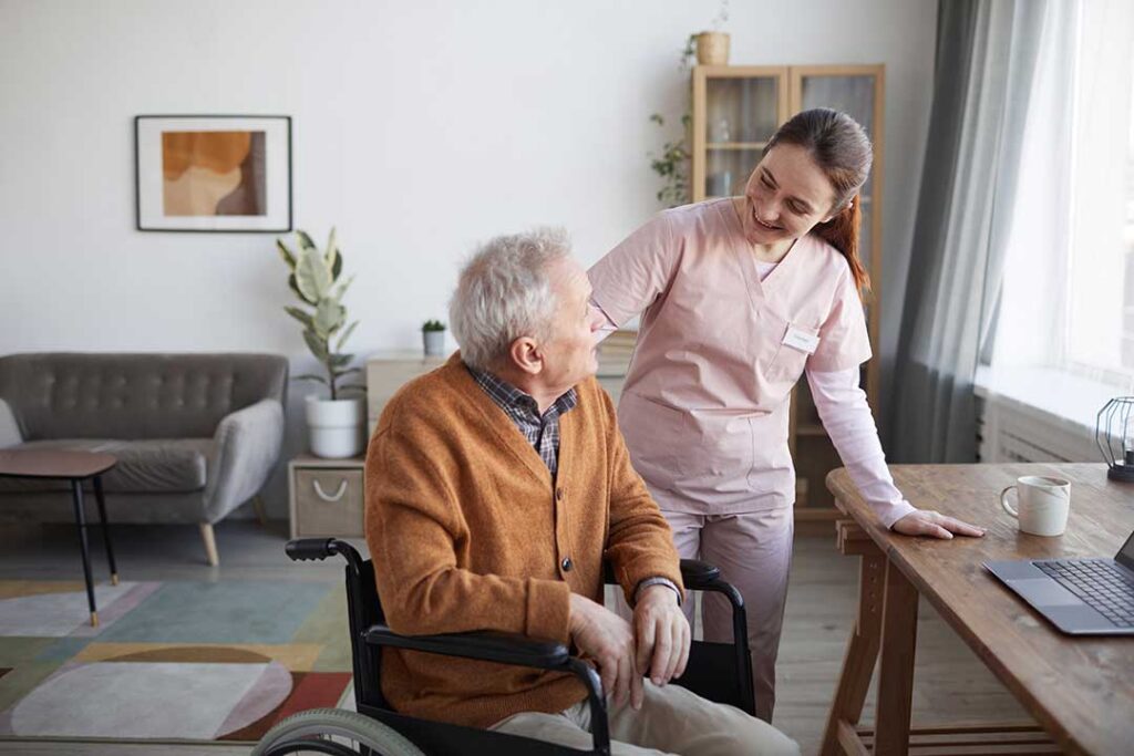 Older resident chats with a caregiver at a small dining table in a bright assisted living common room.