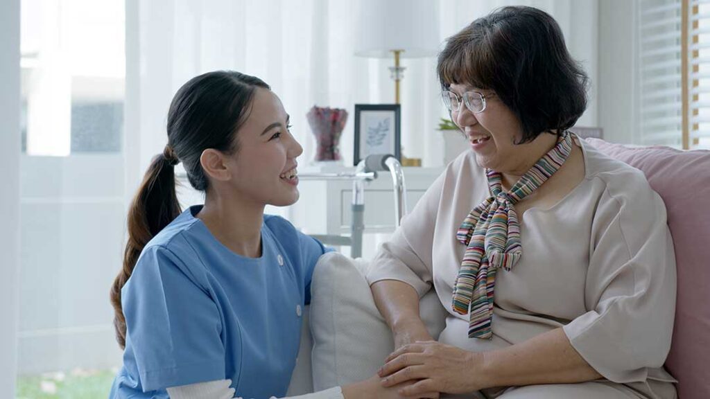 An elderly person and adult family member unpacking boxes together in a sunny, furnished bedroom.